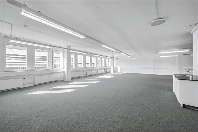 An empty office space with a white ceiling, grey carpet, fluorescent lighting, windows, radiators, and a kitchen with a sink and faucet.