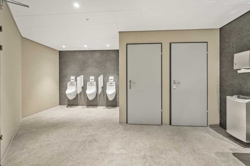 The image shows a commercial bathroom with three urinals mounted on the wall and two gray doors leading to individual stalls. The walls are covered in gray tiles, and the floor is tiled as well. The lighting is bright, creating a clean and modern look.