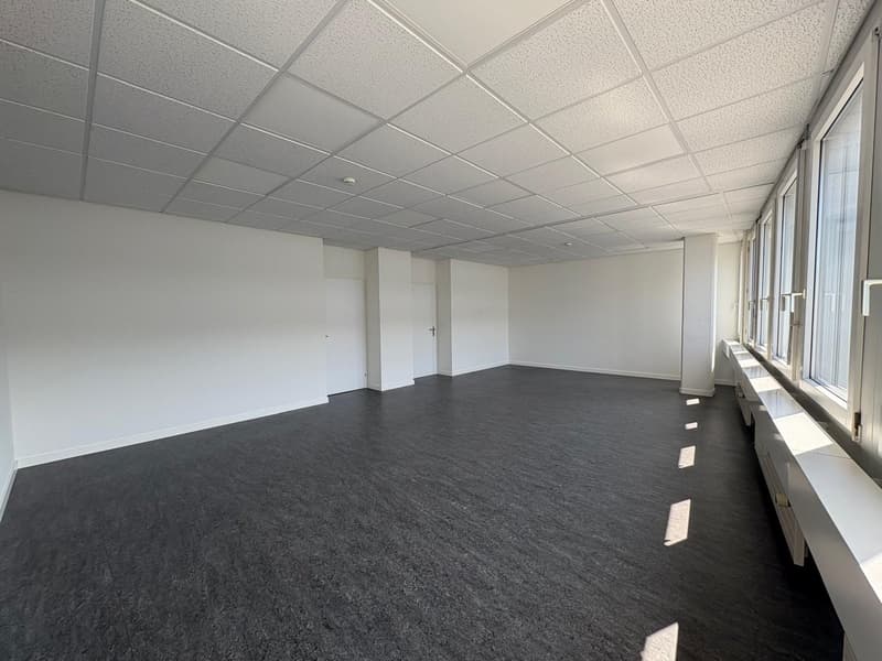 The image shows a large, empty office space with a suspended ceiling and gray carpeted flooring. The room has white walls and large windows along one side, providing natural light. The space appears to be ready for use as an office or commercial space.