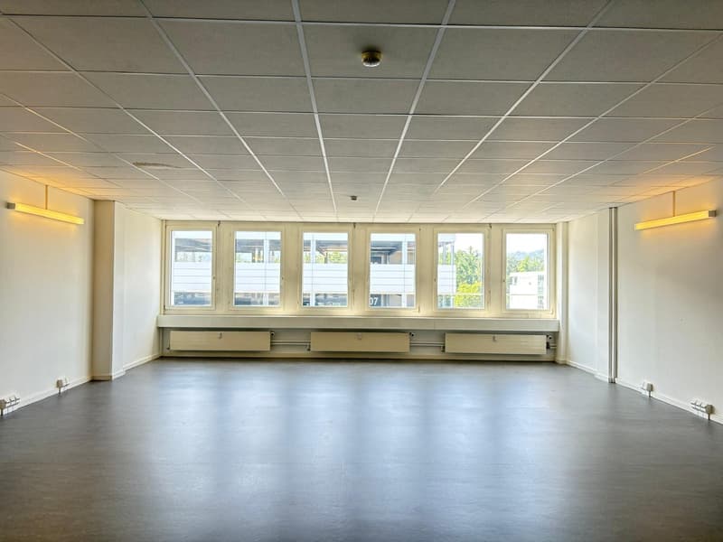 Large open room with a tiled ceiling, multiple windows, and a hardwood floor. The room appears to be an office or meeting space, with no visible furniture or equipment.