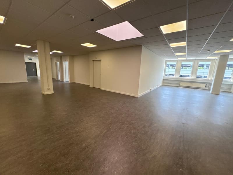 The image shows a large, open office space with a suspended ceiling, recessed lighting, and windows along one wall. The floor is covered in a brown, cork-like material. The space appears to be empty and ready for use or renovation.