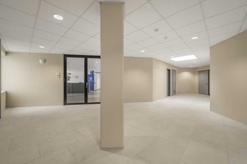 The image shows a large, open office space with a tiled floor, recessed lighting, and a glass-paneled entrance. The space appears to be a commercial or office environment, with a neutral color scheme and minimal furnishings.