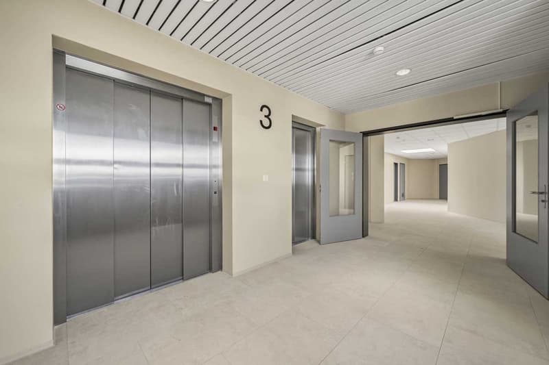 The image shows the interior of a building, with a long hallway and several elevator doors. The hallway has a tiled floor and a white ceiling with recessed lighting. The elevator doors are made of stainless steel and are labeled with the number 3.