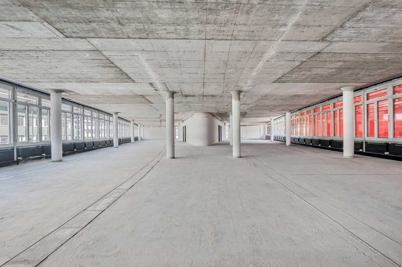 Large open space with concrete floor, high ceiling, and rows of windows along the walls. The space appears to be an unfinished commercial or industrial building, with exposed concrete columns and ceiling.