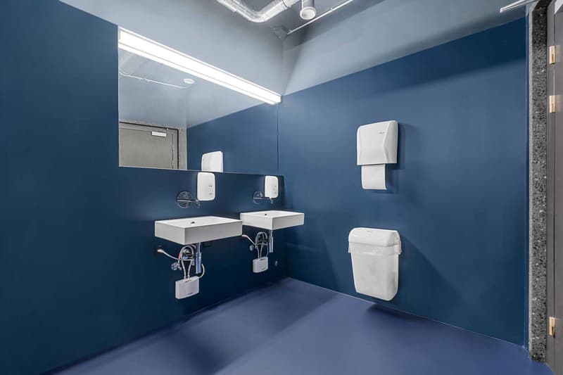 Modern bathroom with blue walls, white fixtures including sinks, soap dispensers, and hand dryers, and a granite or stone countertop.