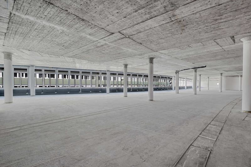 Large open space with concrete floor, exposed ceiling beams, and rows of windows along the walls. The space appears to be a warehouse or industrial building with a minimalist, utilitarian design.