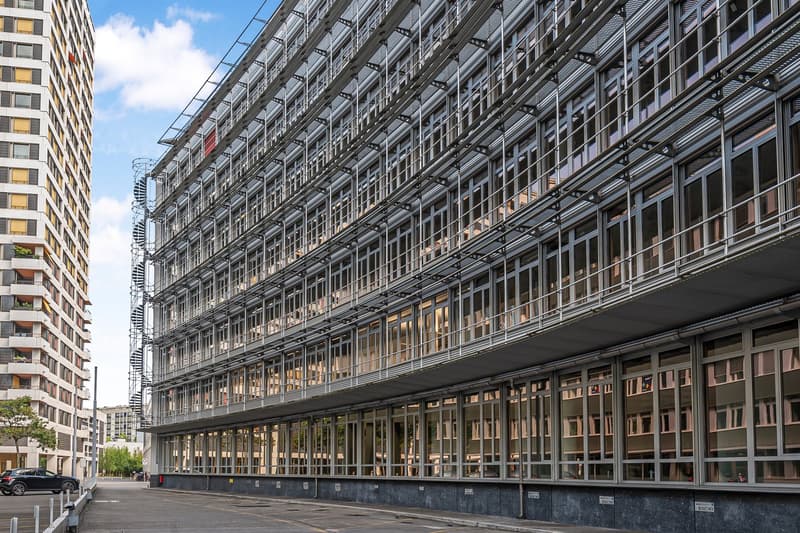 Large multi-story commercial building with a glass and steel facade, featuring a ground-level parking area in front of the building.