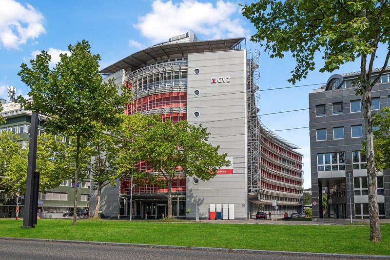 A large modern office building with a curved facade and red accents. The building has multiple floors and is surrounded by trees and greenery.