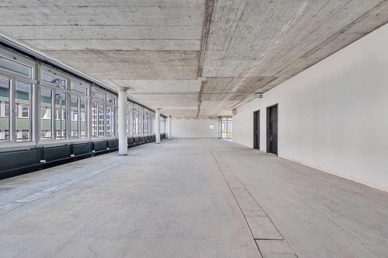 Large open space with concrete floor, high ceiling, and windows along the walls. The space appears to be a commercial or industrial building, possibly an office or warehouse.