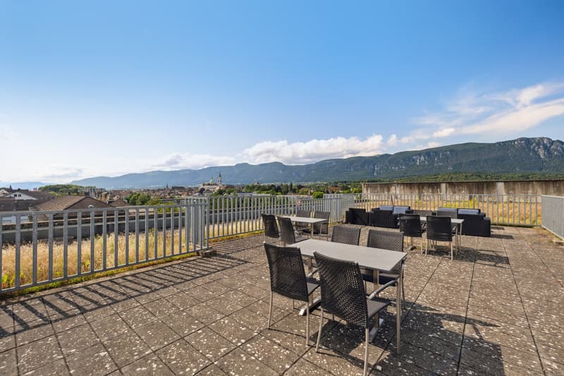 Outdoor seating area with tables and chairs on a rooftop terrace with a view of the town and mountains.