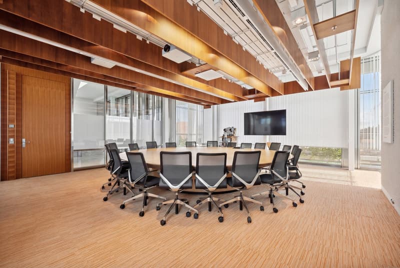 conference room with round table, office chairs, large monitor on wall, wooden ceiling beams, glass walls, wooden entrance door
