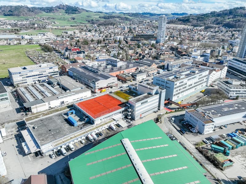 Large industrial area with multiple buildings, some with red tennis courts, multiple solar panels, extensive parking spaces, and multiple cranes and construction vehicles