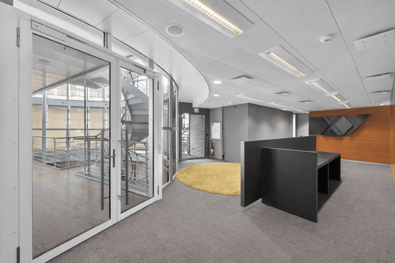 Large open lobby with wide sliding glass doors, a staircase, a desk, and a round carpet. A yellow wall with angular patterns is located to the right side.
