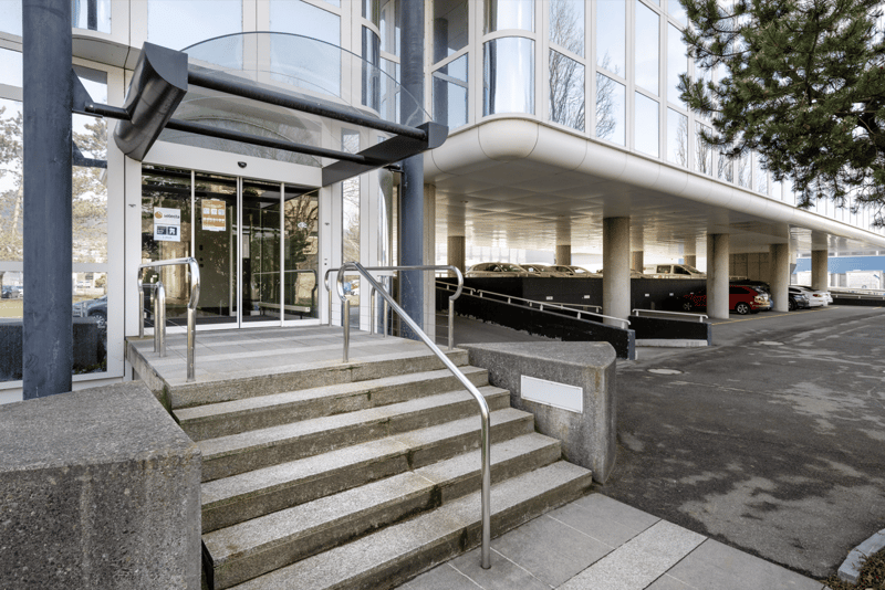 Modern building entrance with glass doors, metal handrails, and a covered walkway. Several cars parked in the parking lot.