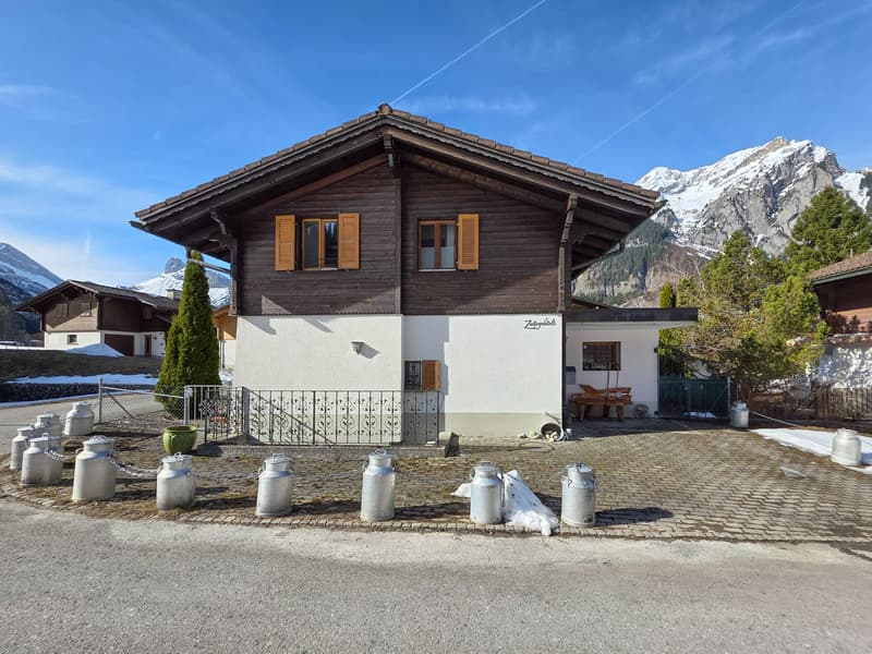 Alpine-style house with wooden exterior, white lower section, brown roof, wooden windows with shutters, surrounded by mountains, milk cans in front