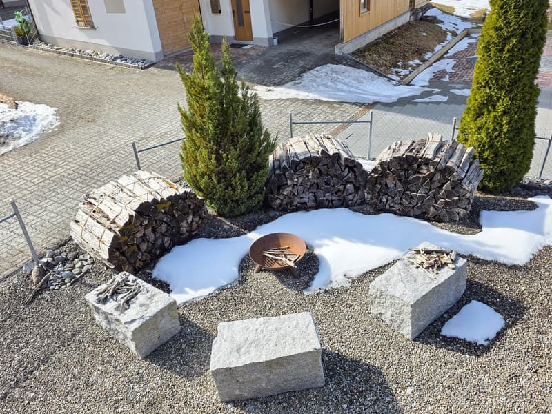 Outdoor fireplace with woodpile, stone seating, snow-covered ground, two evergreen trees, gravel pathway, and a metal fence.