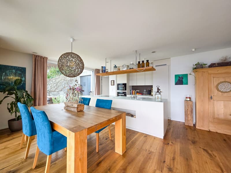 Dining room with wooden table, blue chairs, open kitchen, wooden floor, white walls, large window, wooden cabinet, plant, artwork, hanging lamp