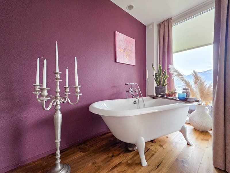 Bathroom with a white bathtub, purple wall, wooden floor, a candle stand with 6 candles, a plant on the window sill, and a window with a view of the mountains.