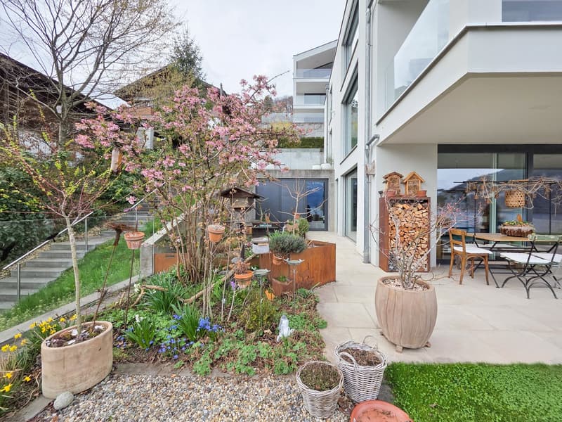 A garden with a variety of plants, trees, and flowers, a patio with a table and chairs, and a building in the background.