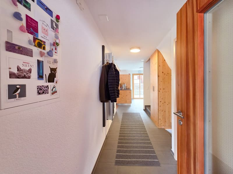 A hallway with a white wall, a wooden door, a rug, and a wooden cabinet with a clock on the wall.