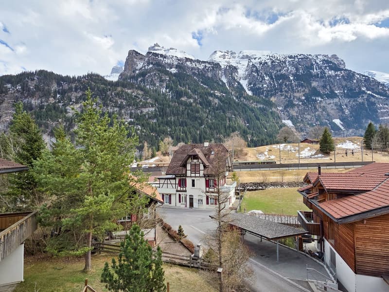 House with brown roof, white walls, red shutters, surrounded by trees and mountains