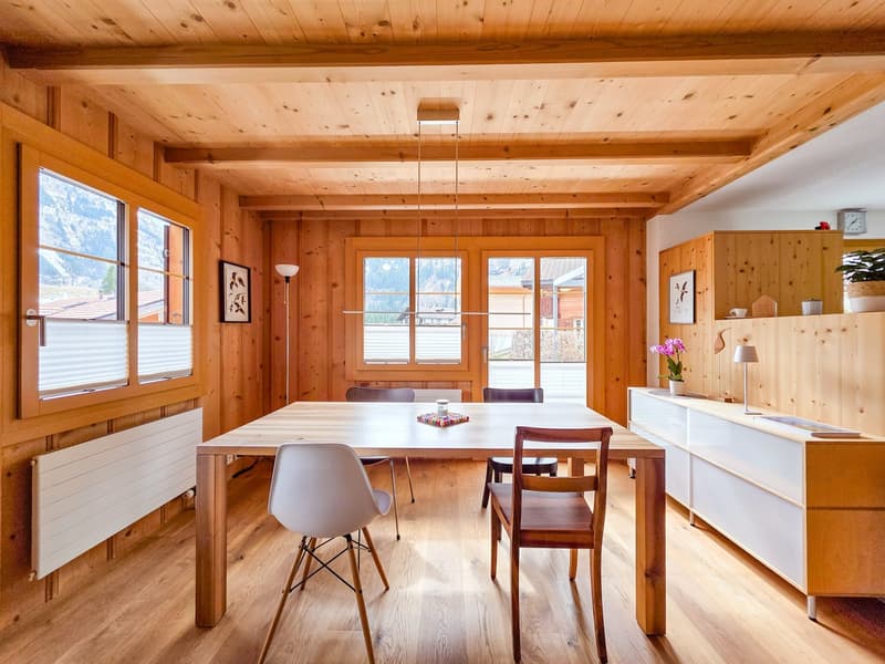 Dining room with wooden walls, wooden floor, large windows, dining table, chairs, radiator, wooden cabinet, flower vase, lamp