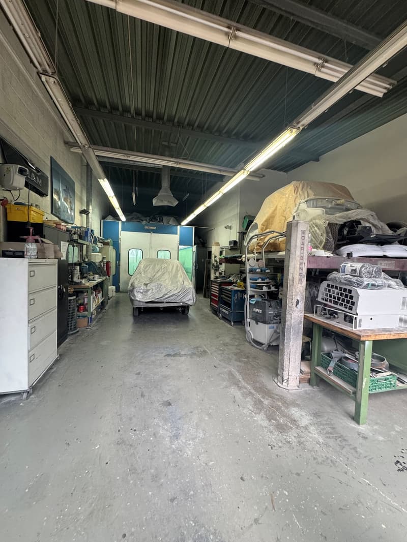 Industrial building with a garage area, metal ceilings, multiple lights, storage shelves, car covered with a tarp, several machines and equipment