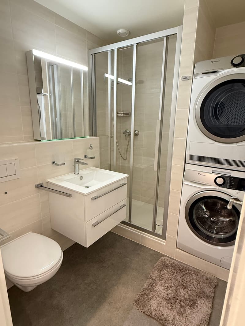 Modern bathroom with white tiles, a glass shower enclosure, a white vanity with a sink, a mirror, a toilet, and a stackable washing machine and dryer.