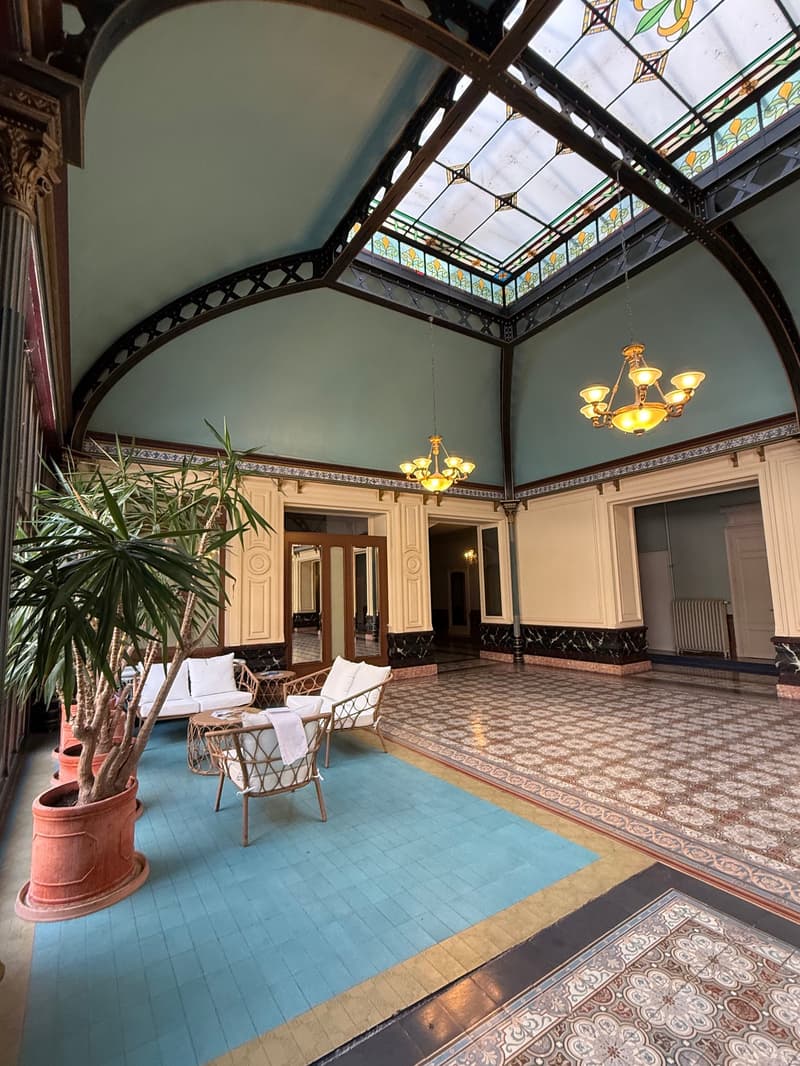 The interior of a building has a high ceiling with stained glass windows, two chandeliers, an ornate wooden ceiling, white walls with decorative border, an arched door, a carpeted floor, a set of white lounge chairs with cushions, a white sofa with cushio