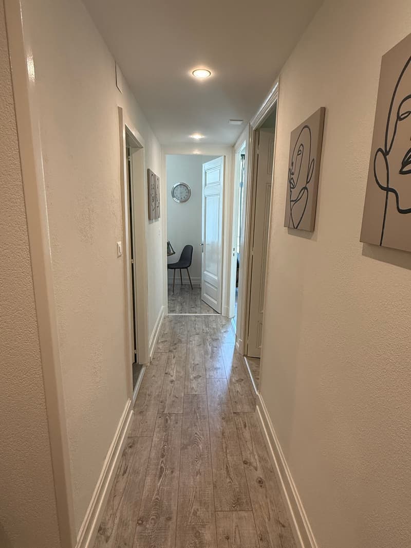 Wide hallway with wooden floors and beige walls, with white doors, a table, and a clock visible through a doorway at the end.