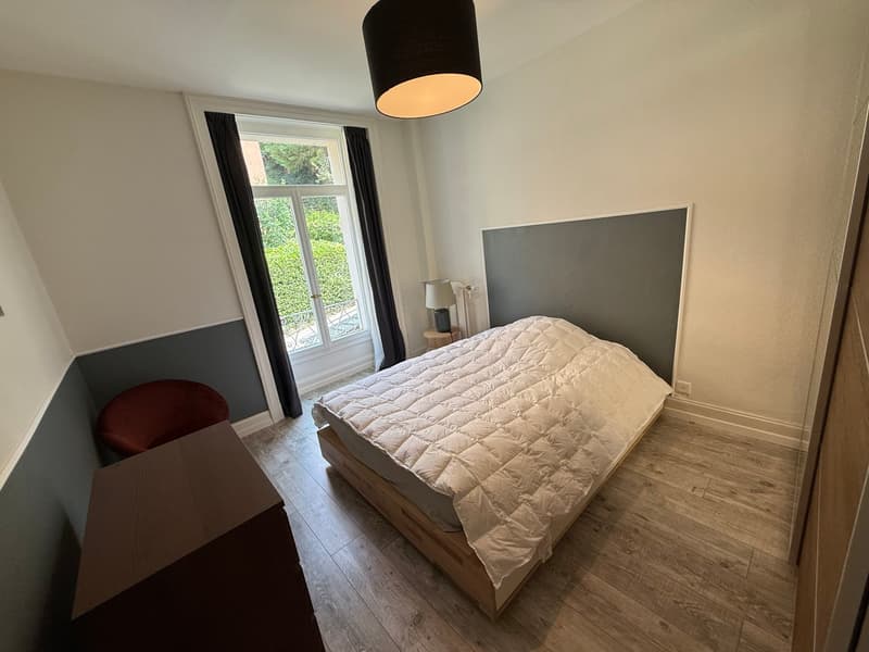 Bedroom with wooden floor, bed, nightstand, lamp, chair, wall-mounted headboard, and a window with curtains.