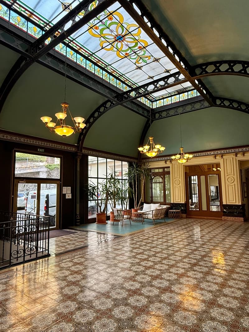 Lobby with stained glass ceiling, two chandeliers, patterned floor, plants in pots, lounge chairs, tables, and arched doorways.