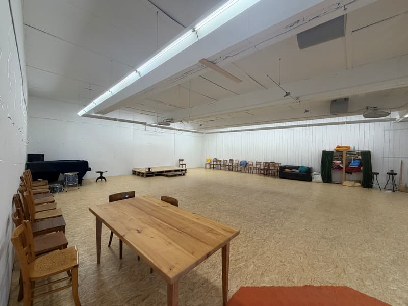 Empty room with wooden table, chairs, and stage, white walls, black piano and drums, ceiling with lights and fan, storage boxes, curtains, and couch.