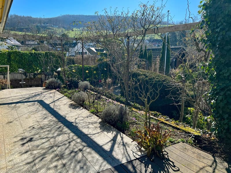 A garden with a wooden deck, a wooden pergola, and a view of the hills and houses in the distance.