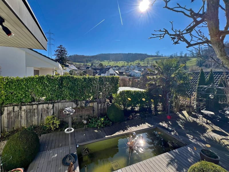 Wooden deck, small pond, garden with various plants, hedges, and trees, distant houses and hills