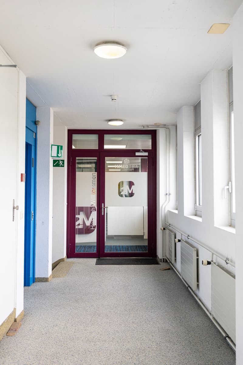 The image shows a hallway with a burgundy-colored glass door leading to an office or business. The hallway has white walls, a tiled floor, and ceiling lights. There are emergency exit signs and other signage visible on the walls.
