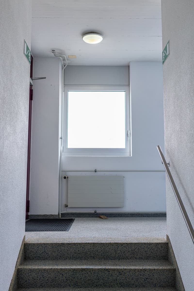 The image shows a stairwell with a window at the top, a radiator on the wall, and a textured floor with a mat at the bottom of the stairs.