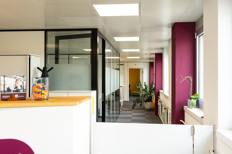 Modern office space with glass partitions, wooden countertop, and colorful decor elements. The space features a reception area, hallway with glass walls, and a mix of open and private work areas.