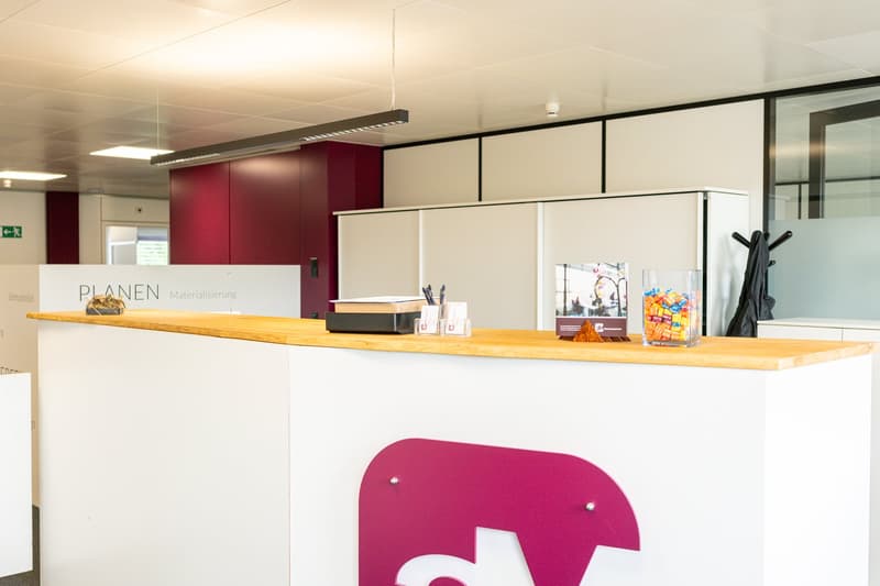The image shows an office space with a long wooden counter, a pink elephant-shaped decoration, and various office supplies and equipment on the counter. The space has white walls and a red accent wall, with a sign that says 'PLANEN Materialisierung' (whic
