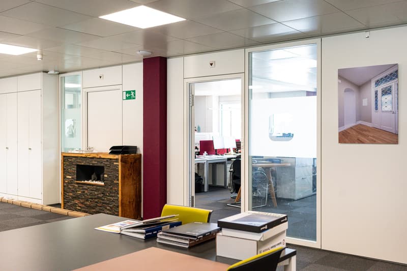 The image shows an office space with a fireplace, storage cabinets, and various office equipment and supplies. The space has a modern and professional design with white walls, tiled floors, and recessed lighting. There is a glass door leading to another r