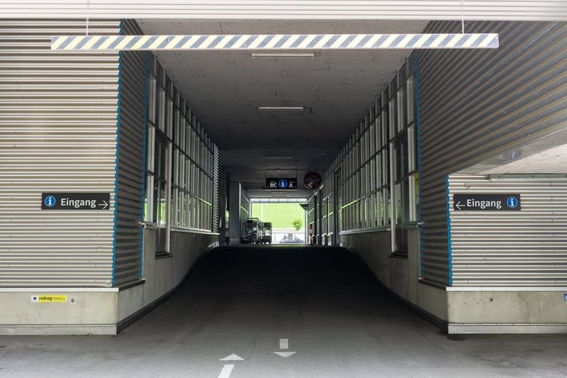 A long, enclosed walkway or passage with concrete walls and a striped ceiling, leading to an open outdoor area with a grassy field visible at the end. The passage has signs indicating the entrance on both sides.
