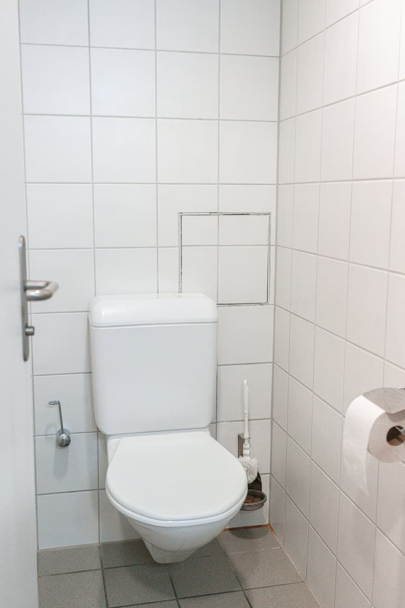 The image shows a bathroom with white tiled walls, a white toilet, and a small shelf or storage space next to the toilet.