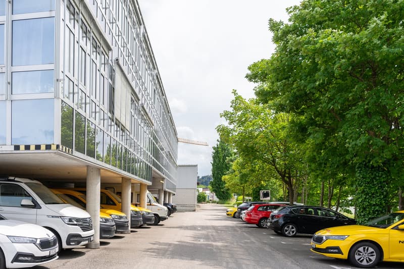 A multi-story commercial building with a glass facade, surrounded by a parking lot with various cars parked, including yellow taxis. The building is situated in a green, tree-lined area.