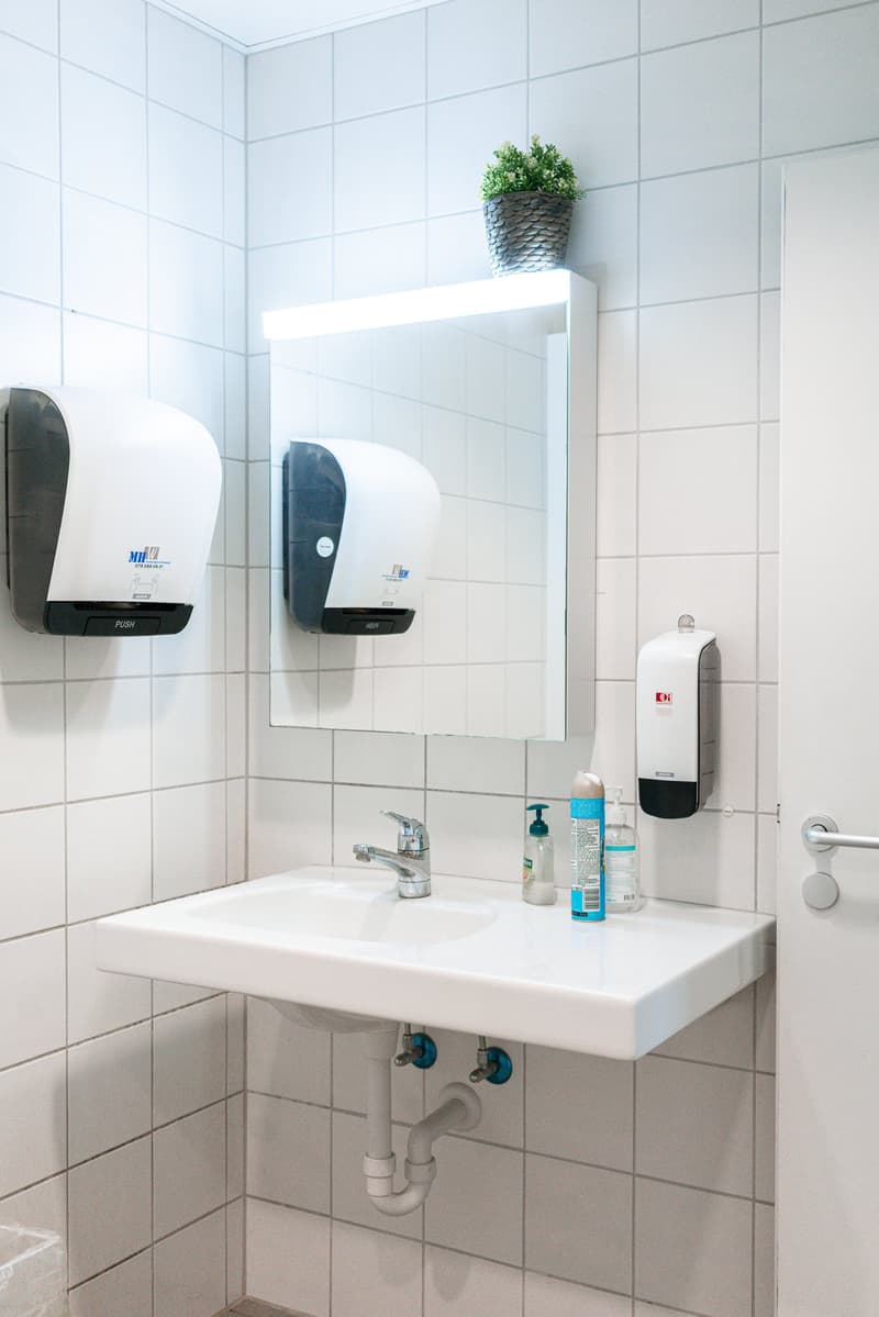 White tiled walls, white sink with faucet, wall-mounted soap dispenser, two hand dryers, and a potted plant on top of the mirror cabinet