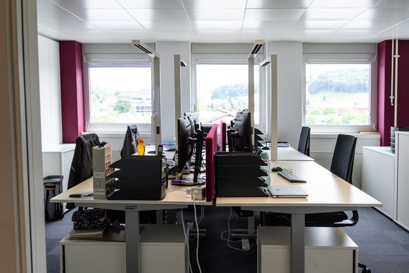 The image shows an office space with multiple desks and computer workstations. The room has large windows providing natural light, and the walls are painted in a light gray color. There are several black office chairs and the desks are cluttered with vari