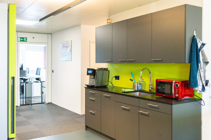 The image shows a small office kitchen area with dark gray cabinets, a green backsplash, and various kitchen appliances such as a microwave, coffee maker, and a red toaster oven. There is also a sink and a dishwasher visible. The kitchen is located in an