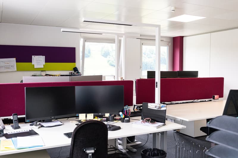 The image shows an office space with multiple workstations. The room has white walls, a purple and green accent wall, and large windows providing natural light. The workstations have multiple computer monitors, keyboards, and other office equipment. The f