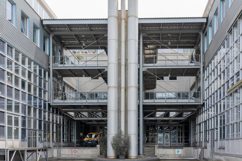 The image shows a large commercial building with a modern, industrial-style architecture. The building has a glass facade with multiple floors and balconies. There are large columns supporting the structure, and a yellow car can be seen parked in the fore