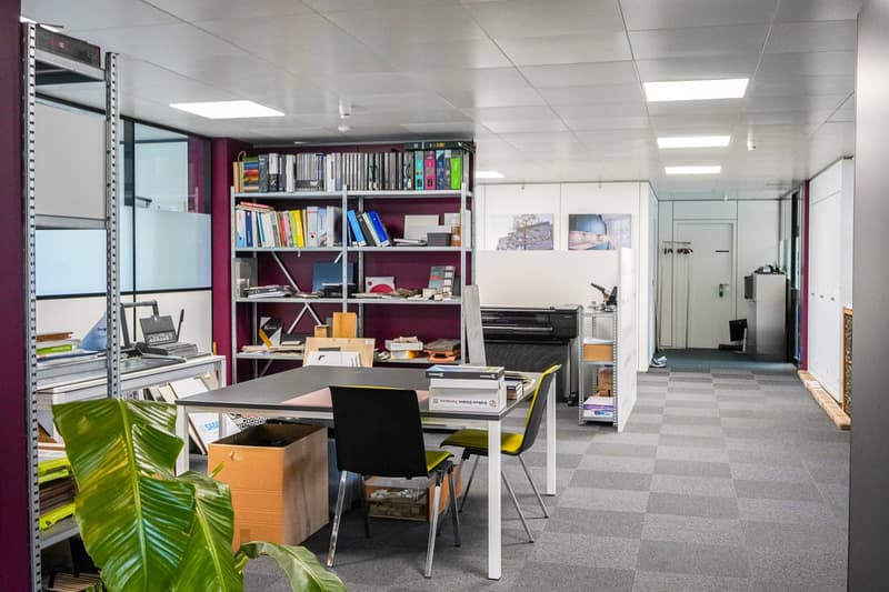 The image shows an office space with a desk, computer, and various office supplies and equipment. The room has shelves filled with books and other items, and the walls are decorated with artwork. The space appears to be organized and functional, with a mi