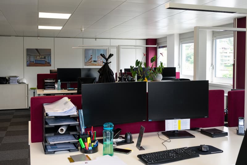 The image shows an office space with multiple workstations. The office has white walls, a suspended ceiling with recessed lighting, and large windows providing natural light. The workstations are equipped with desktop computers, monitors, keyboards, and o